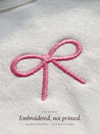 Close-up of the pink satin-stitch coquette bow embroidery on a white crewneck