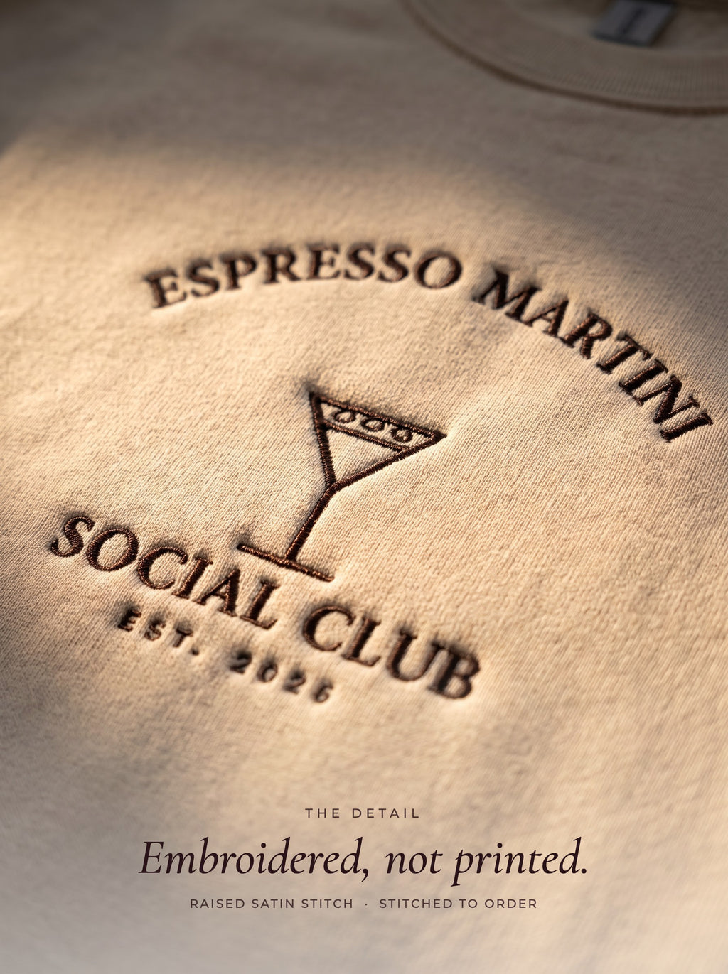 Close-up of the Espresso Martini Social Club embroidery in raised dark brown thread on a sand crewneck