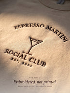 Close-up of the Espresso Martini Social Club embroidery in raised dark brown thread on a sand crewneck
