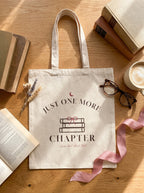 Natural canvas Just One More Chapter tote bag with stacked books and pink ribbon on a wooden table
