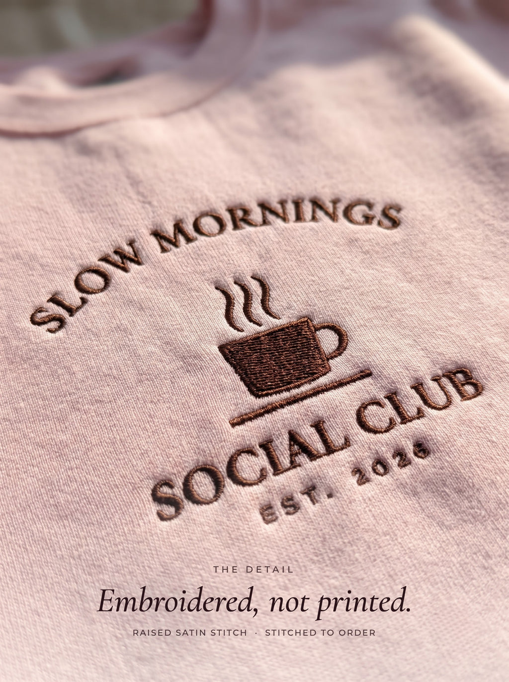 Close-up of the Slow Mornings Social Club embroidery in raised dark brown thread on a light pink crewneck