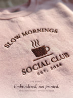Close-up of the Slow Mornings Social Club embroidery in raised dark brown thread on a light pink crewneck