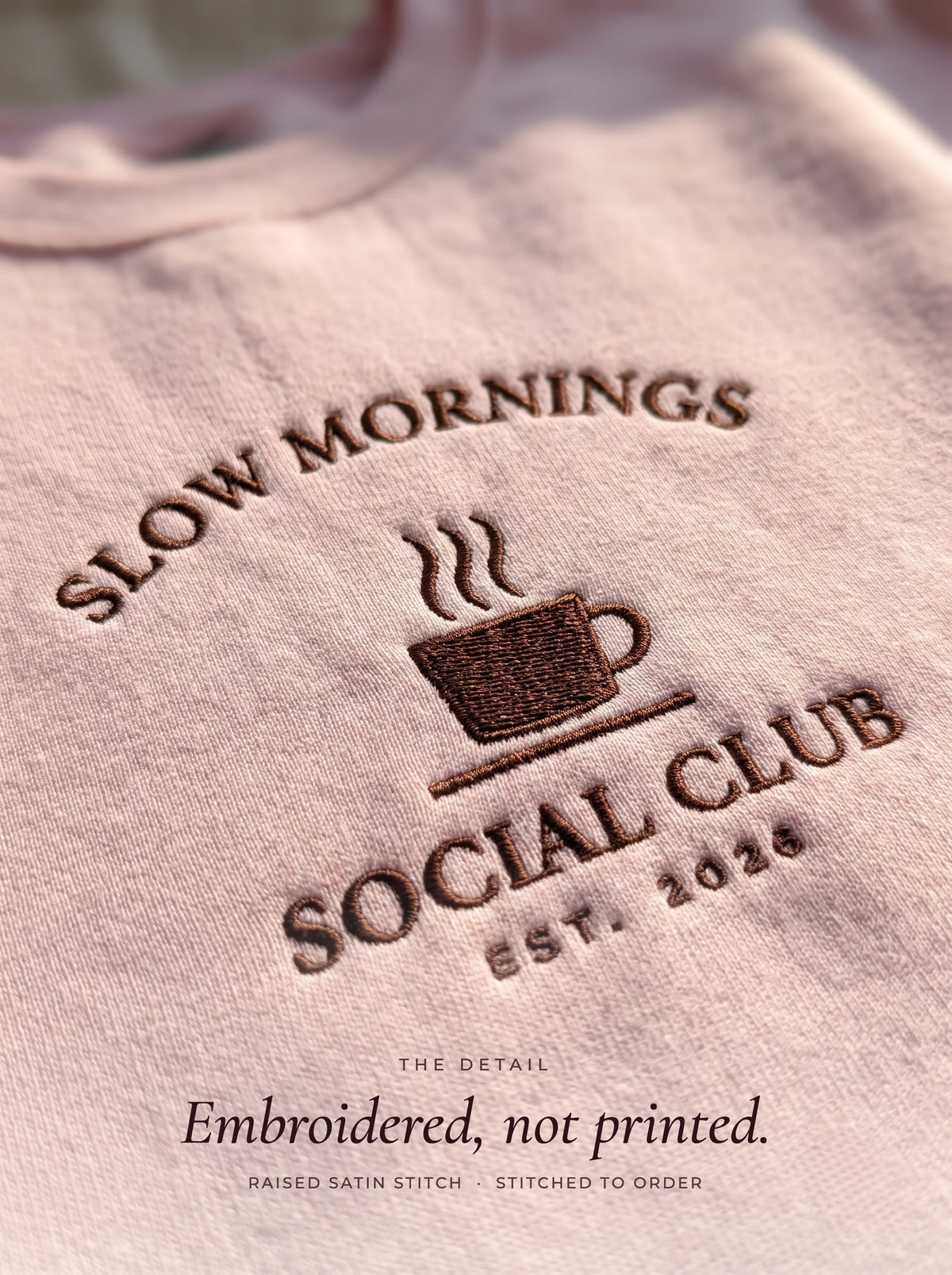Close-up of the Slow Mornings Social Club embroidery in raised dark brown thread on a light pink crewneck