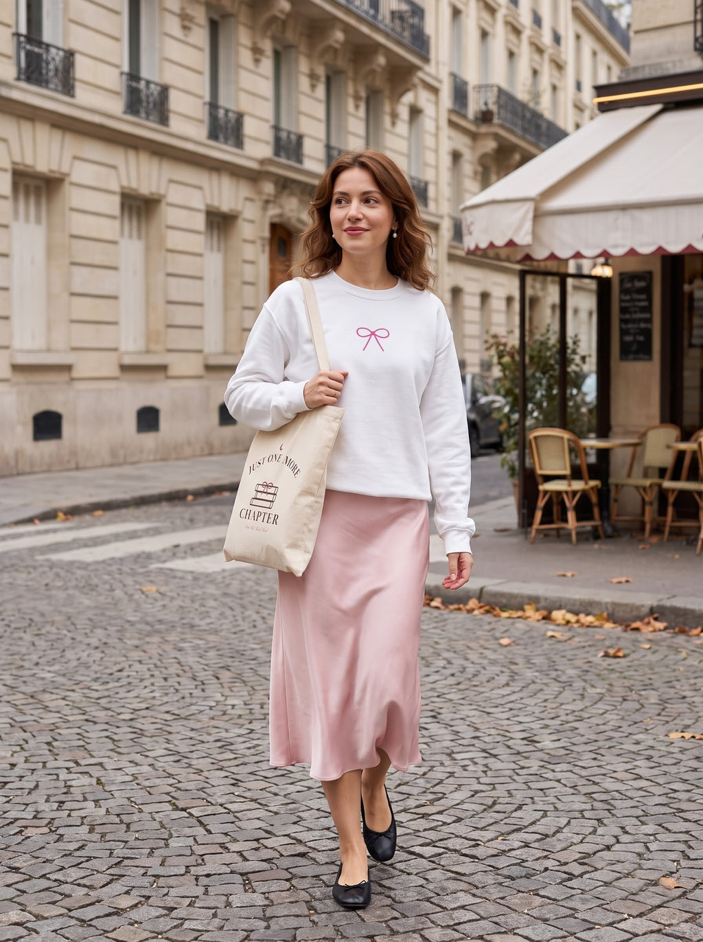 Smiling woman in the white Coquette Bow sweatshirt and pink satin midi skirt outside a Paris café