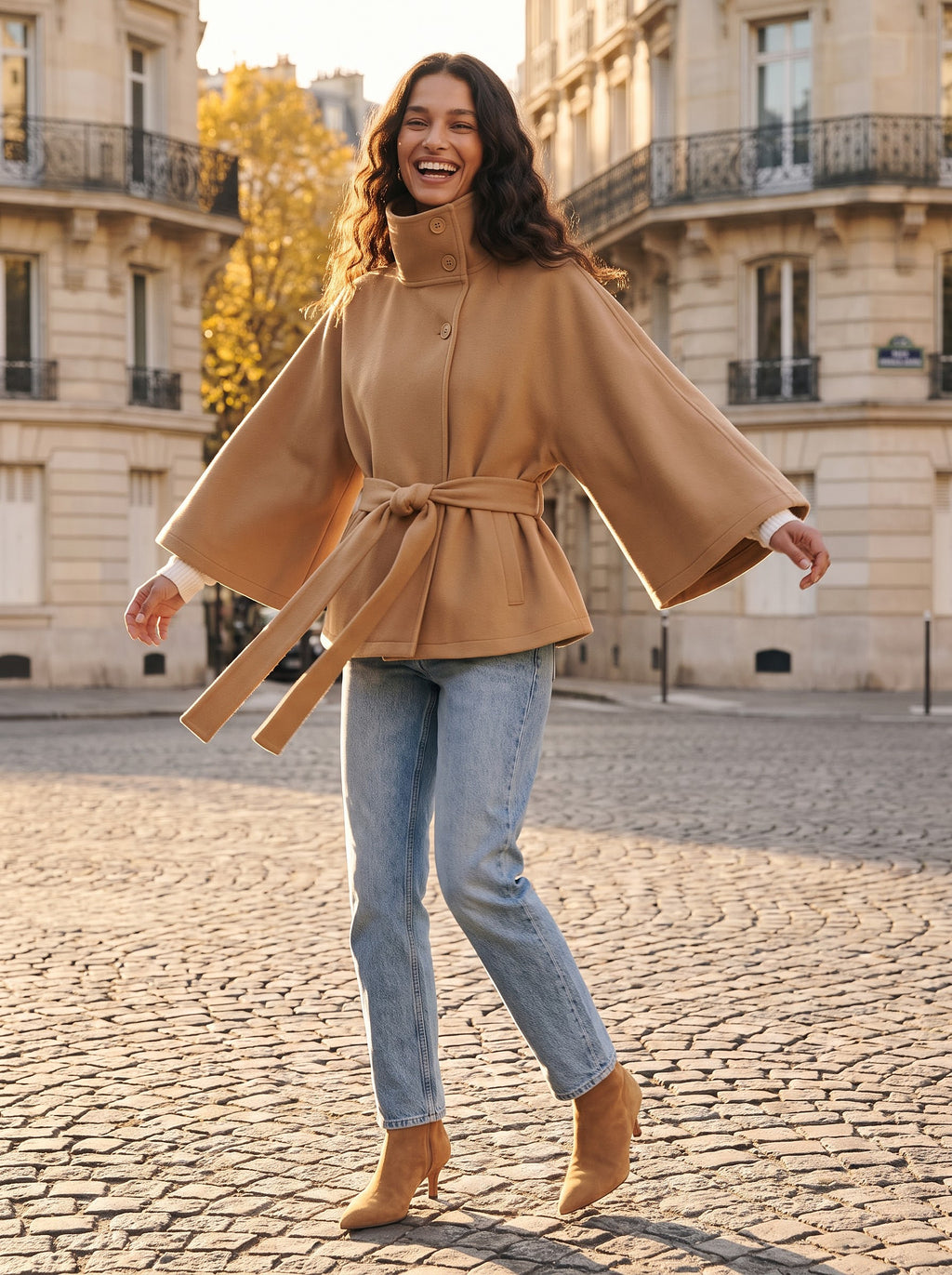 Model spinning on a Paris cobblestone street so the camel cape sleeves and tie belt of the City Muse coat swing out