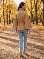 Back view of the camel City Muse cape coat with tie belt, worn on an autumn park path