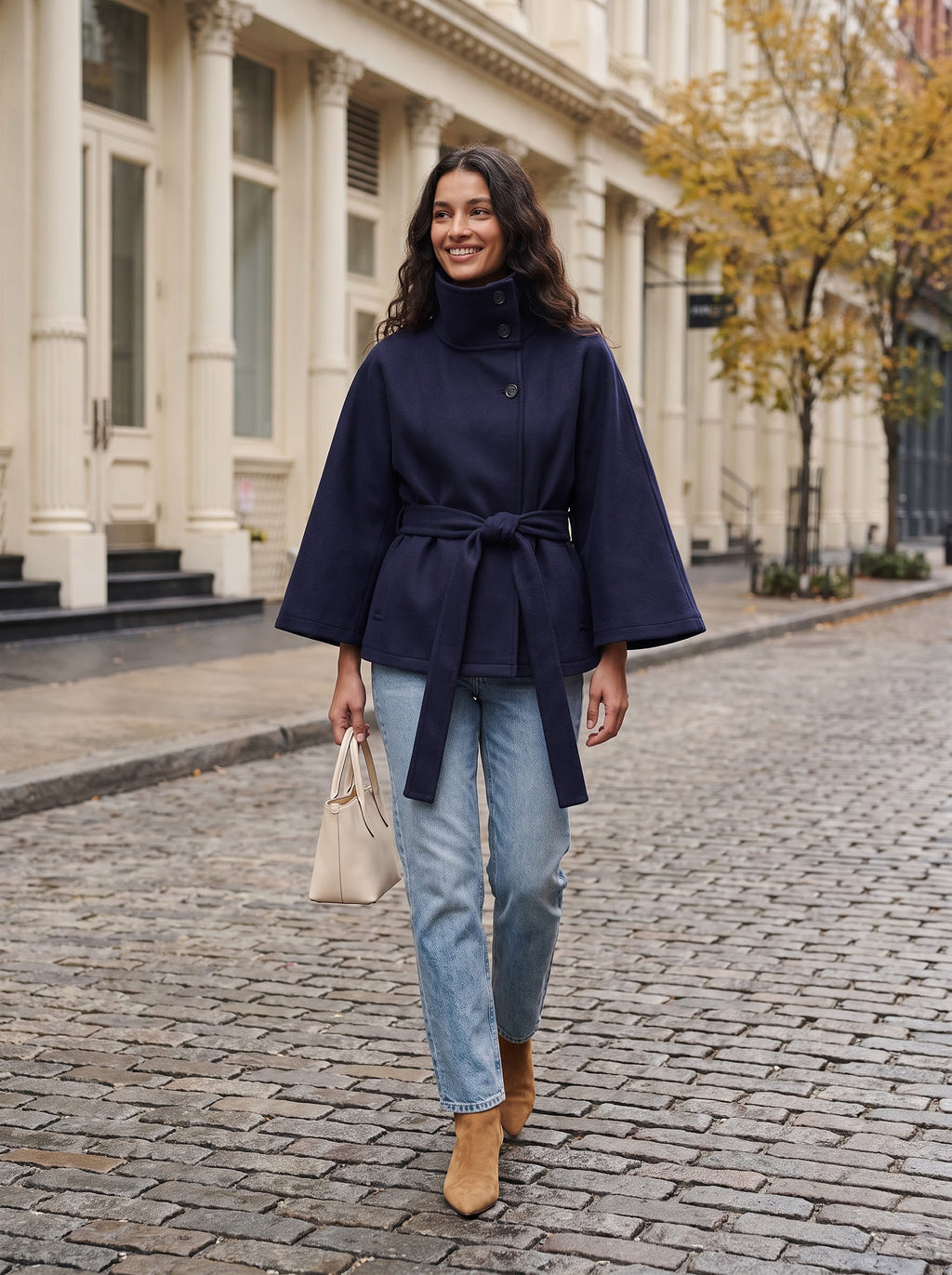 Woman walking on a SoHo cobblestone street in the navy City Muse cape coat with a cream tote bag