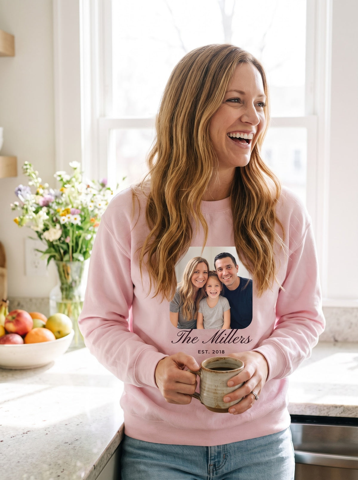 Woman holding a coffee mug in a sunny kitchen, wearing a light pink custom family photo crewneck sweatshirt