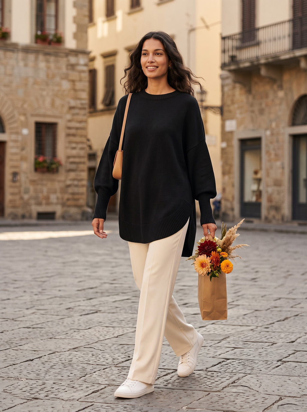 Woman walking across a city plaza in the black oversized tunic sweater with cream wide-leg trousers and sneakers