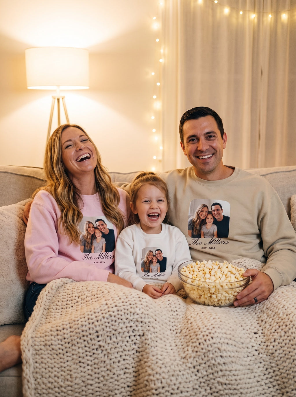 Family of three cuddling on the couch on movie night in matching custom photo crewneck sweatshirts in light pink, sand and white