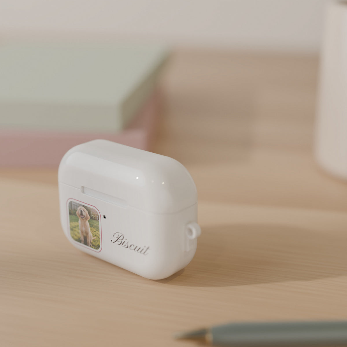 Custom photo AirPods Pro case with a golden retriever photo and the name Biscuit in script, standing on a light oak desk beside a mug, books and a pen