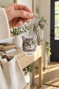 Acrylic cat-head keychain with a white tab printed Luna in script, on a chain and split ring with keys, held in a hand in a sunny entryway