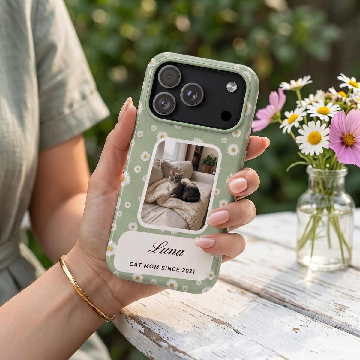 Sage daisy phone case with a cat photo window, held over a garden table with fresh daisies