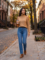 Woman walking on an autumn city street in the khaki camel cold-shoulder turtleneck sweater with jeans, ankle boots and a coffee cup