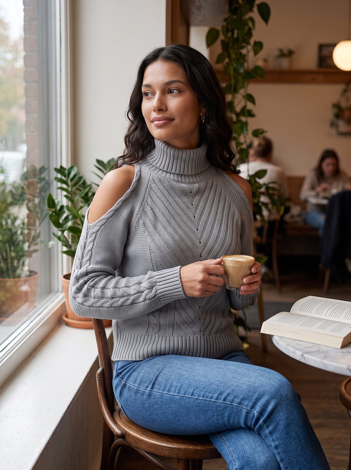Woman sitting by a cafe window in the gray cold-shoulder turtleneck sweater holding a latte
