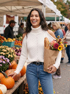 Woman at an autumn farmers market wearing the white cold-shoulder turtleneck sweater with jeans, holding flowers and a baguette