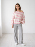 Weekend Stripe off-shoulder knit sweater in pink and cream wide stripes, worn with gray wide-leg trousers, studio photo